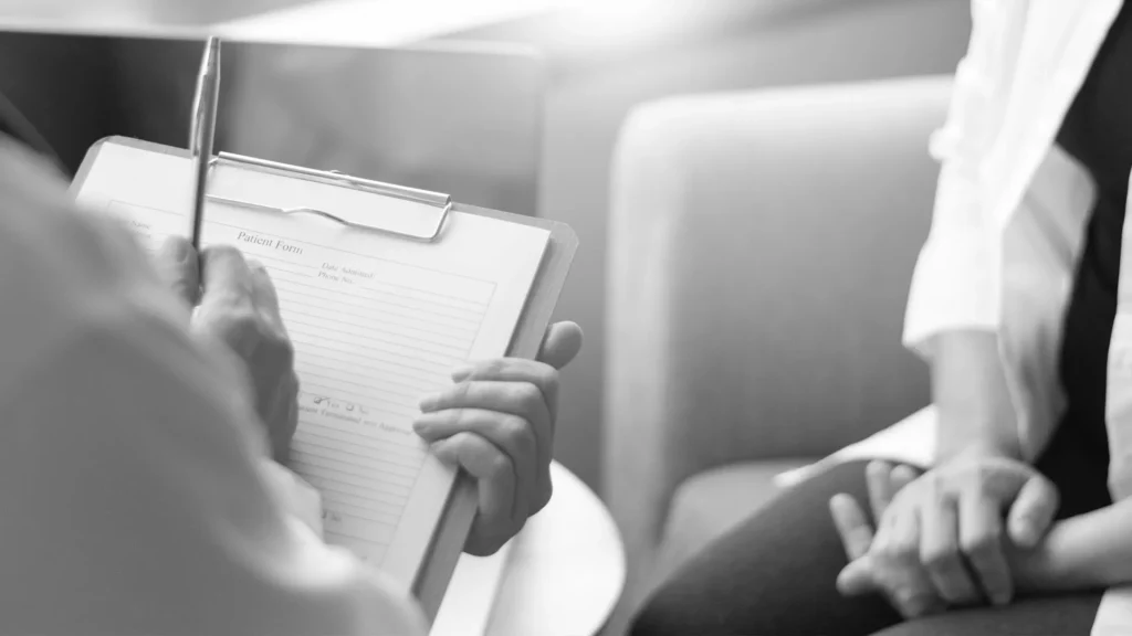 Person hält Klemmbrett mit Patientenformular und schreibt mit Stift, gegenüber sitzt eine Person mit gefalteten Händen im Gesprächskontext